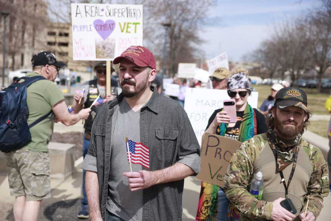 Two younger men are shown. One is wearing a t-shirt with camouflage print; the other carries a small American flag. 