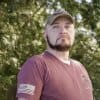 Kevin Conlon wears a dark red t-shirt with an American flag on it and an army green baseball cap. 