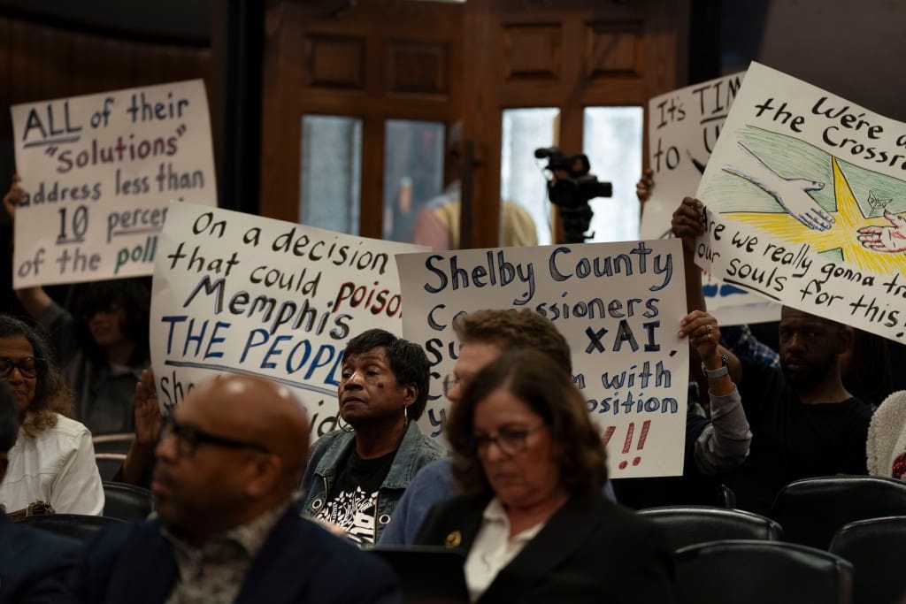A Black woman sits at a meeting in front of a row of signs held protesting xAI.